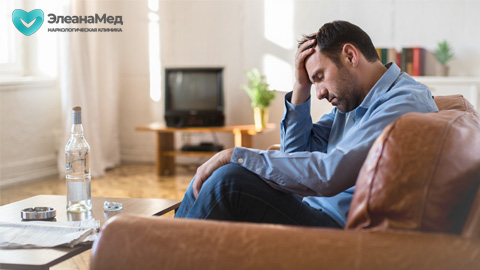 Мужчина сидит на диване с бутылкой алкоголя, держится за голову